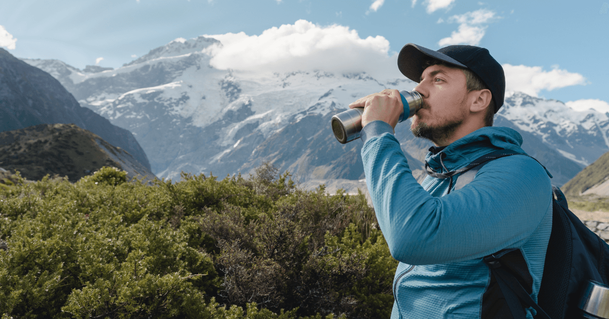 A man drinks water while standing before a majestic mountain range, showcasing nature's beauty and tranquility.
