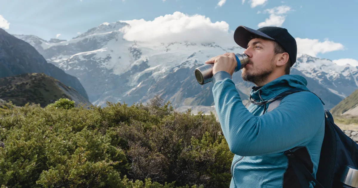 A man drinks water while standing before a majestic mountain range, showcasing nature's beauty and tranquility.
