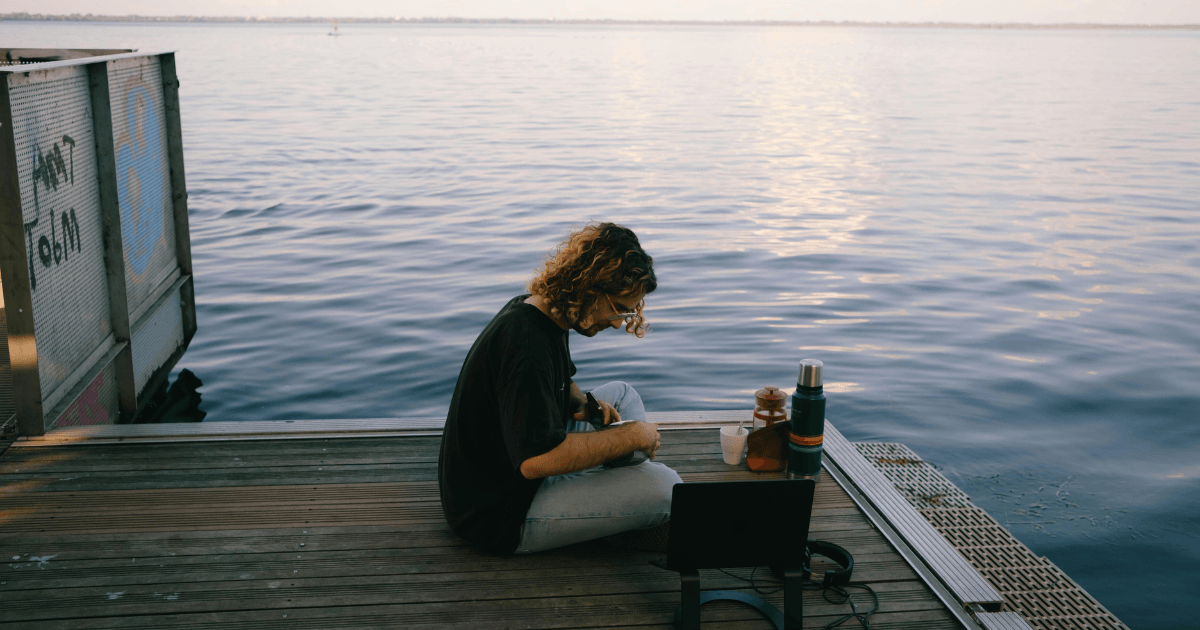 A person seated on a wooden dock, gazing out over a tranquil body of water under a clear blue sky.