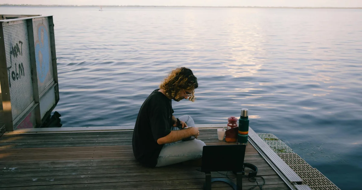 A person seated on a wooden dock, gazing out over a tranquil body of water under a clear blue sky.