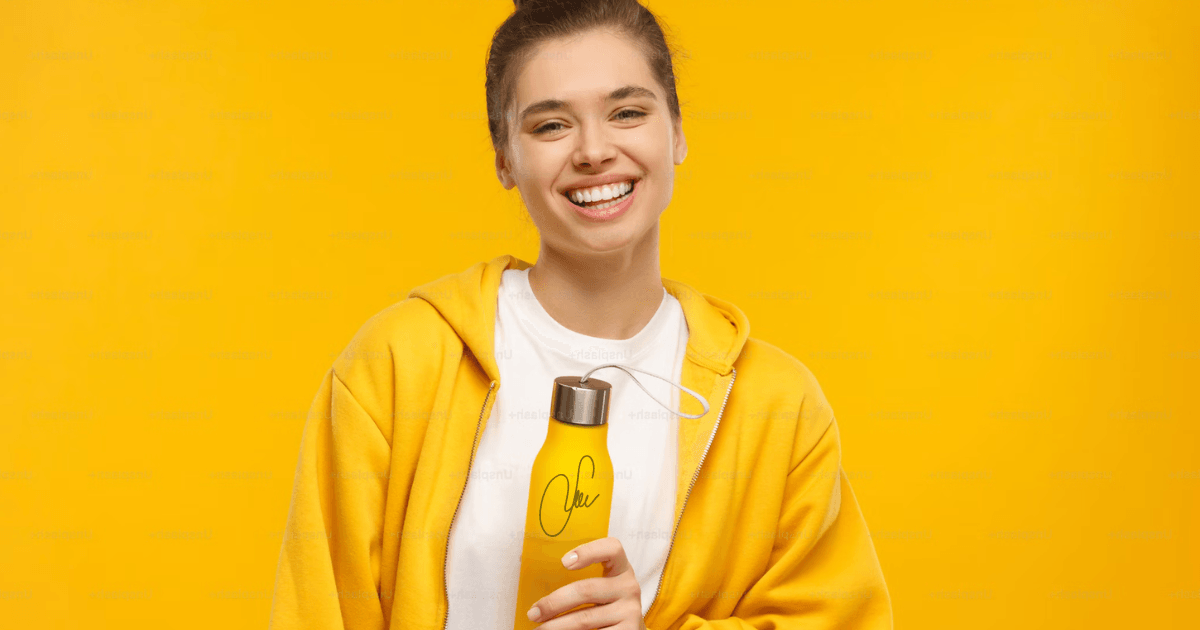 A woman wearing a yellow hoodie is holding a bottle of water, smiling in a bright outdoor setting.