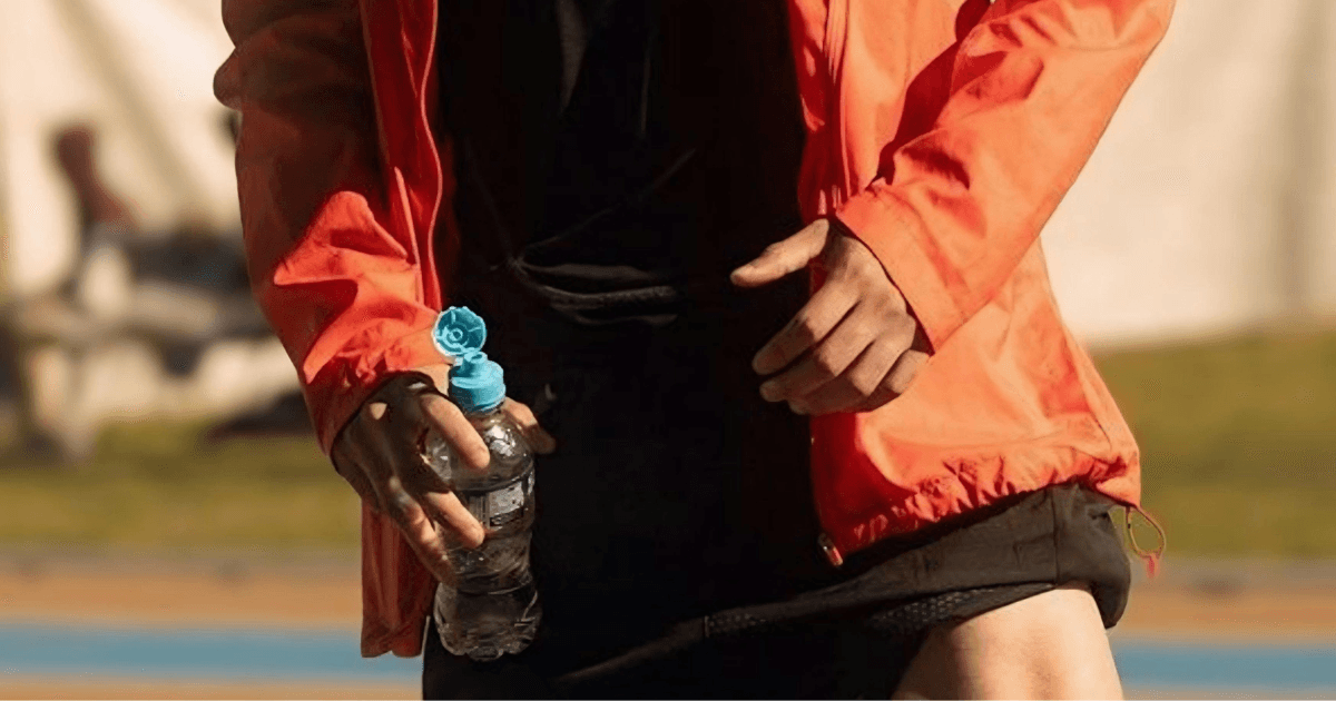 A man wearing an orange jacket is holding a water bottle in his right hand, standing outdoors.
