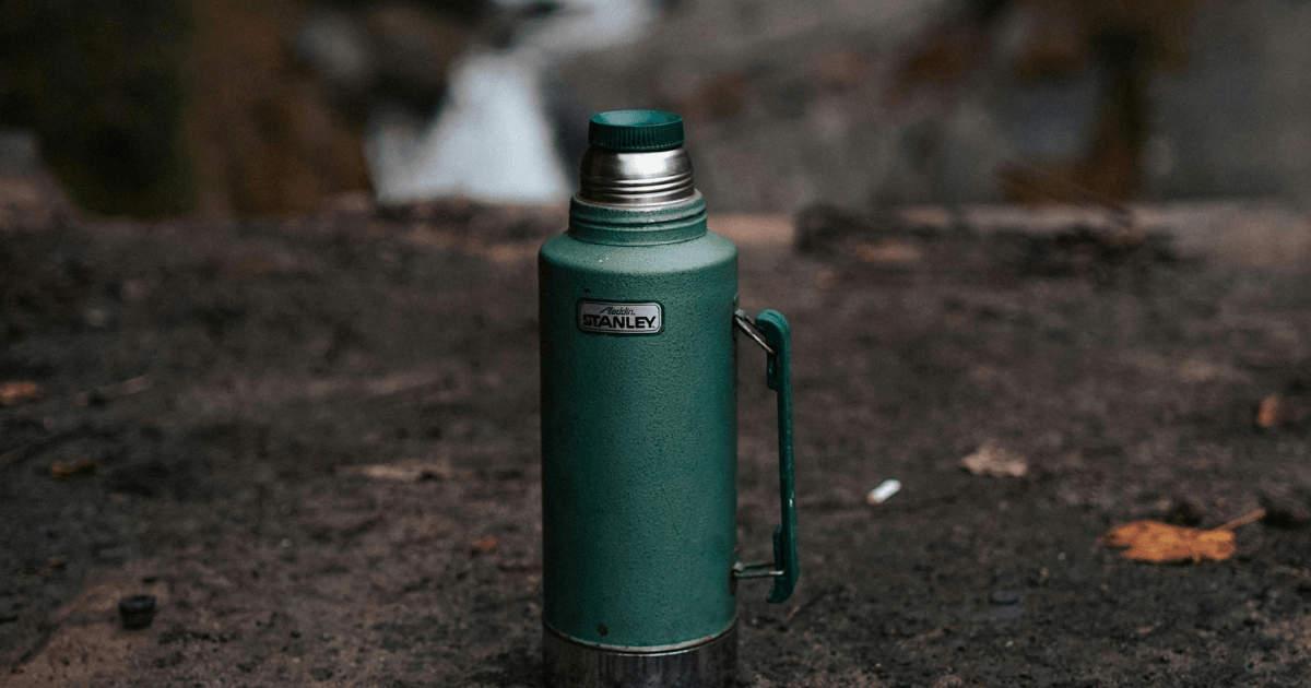 A green water bottle rests on the ground beside a flowing waterfall, surrounded by natural beauty.