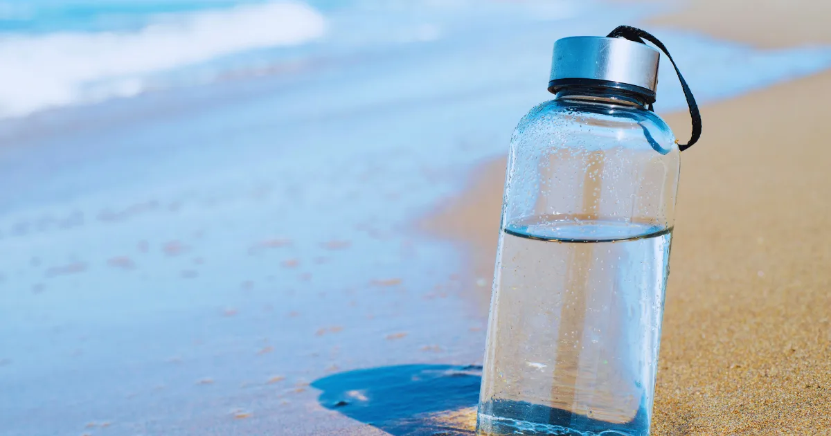 A water bottle rests on the sandy beach, with the ocean waves gently lapping in the background.