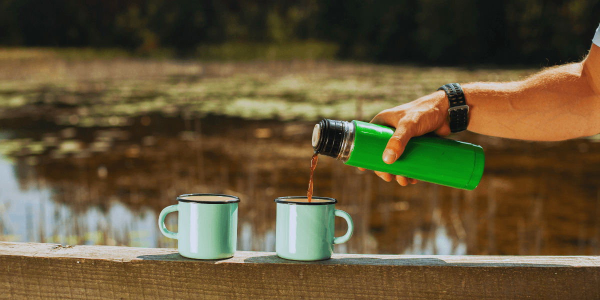 A man pours coffee into two cups while standing on a wooden bridge, surrounded by a serene natural setting.