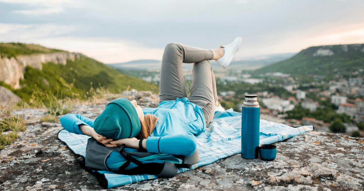 A woman relaxes on a blanket, her feet elevated, surrounded by a scenic mountain landscape.