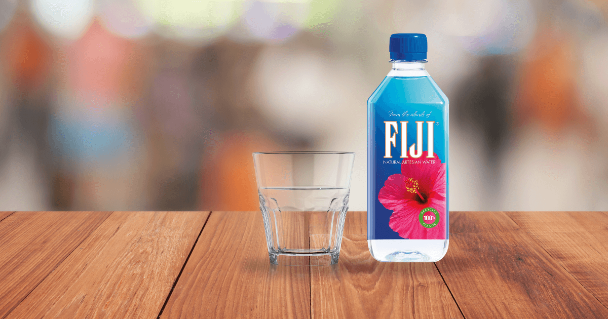 A clear glass and a 500ml bottle of FIJI Natural Artesian Water placed on a polished wooden table.