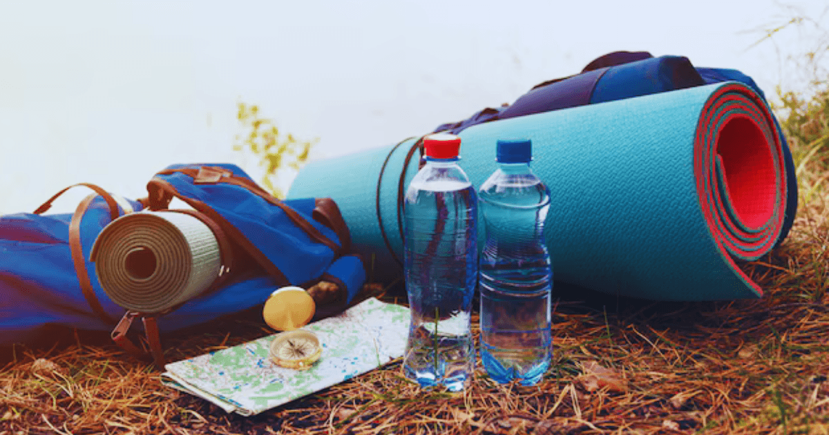 A camping setup with two plastic water bottles, a rolled-up turquoise yoga mat with red edges, and a blue backpack with leather straps.