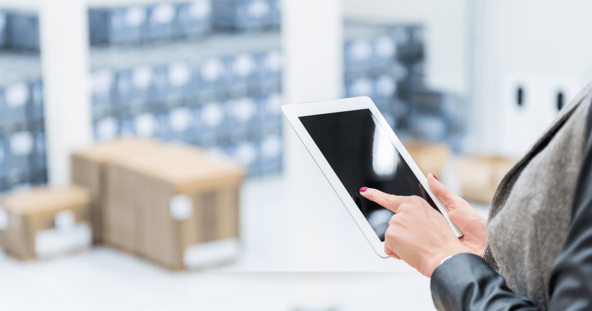 A woman stands in a warehouse, holding a tablet computer while reviewing inventory or managing logistics.