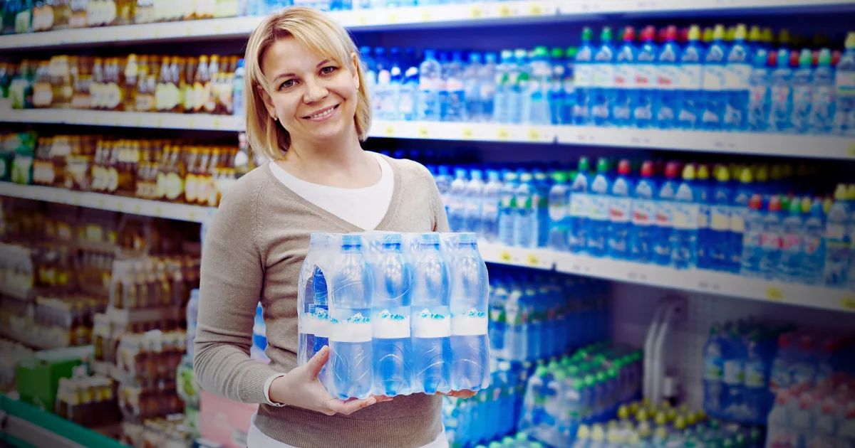 A woman holding a clear bottle of water, smiling and standing outdoors in a sunny environment.