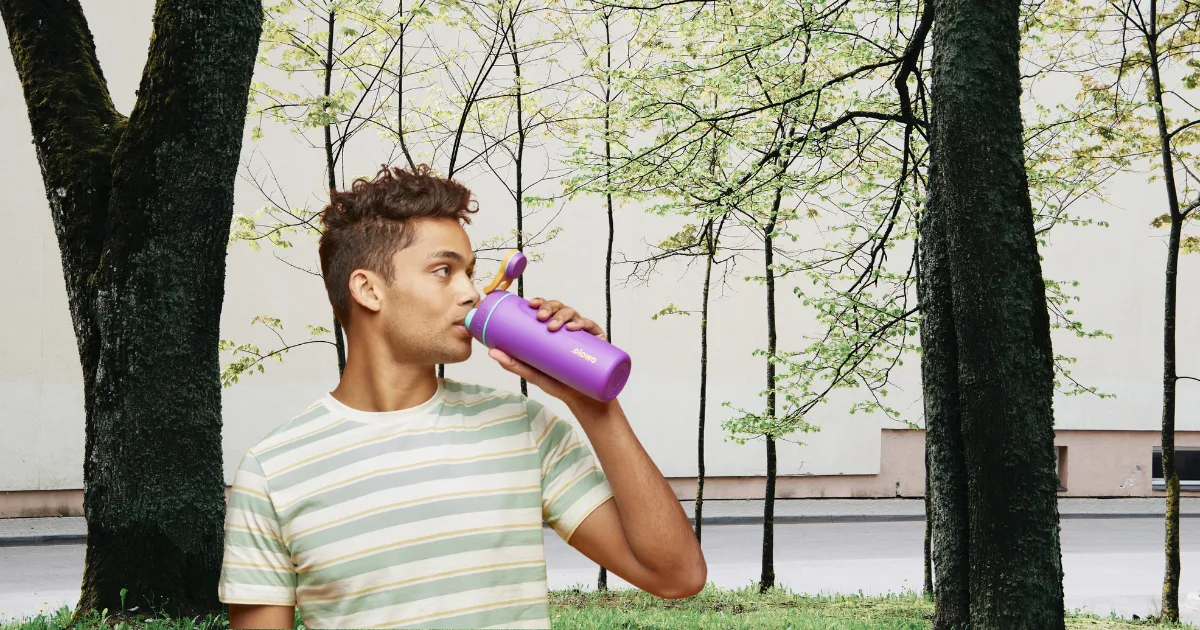 A person wearing a striped shirt drinks from an Owala water bottle, the perfect hydration solution, while standing between two large trees. The background features a wall, leaf-covered trees, and a paved path.