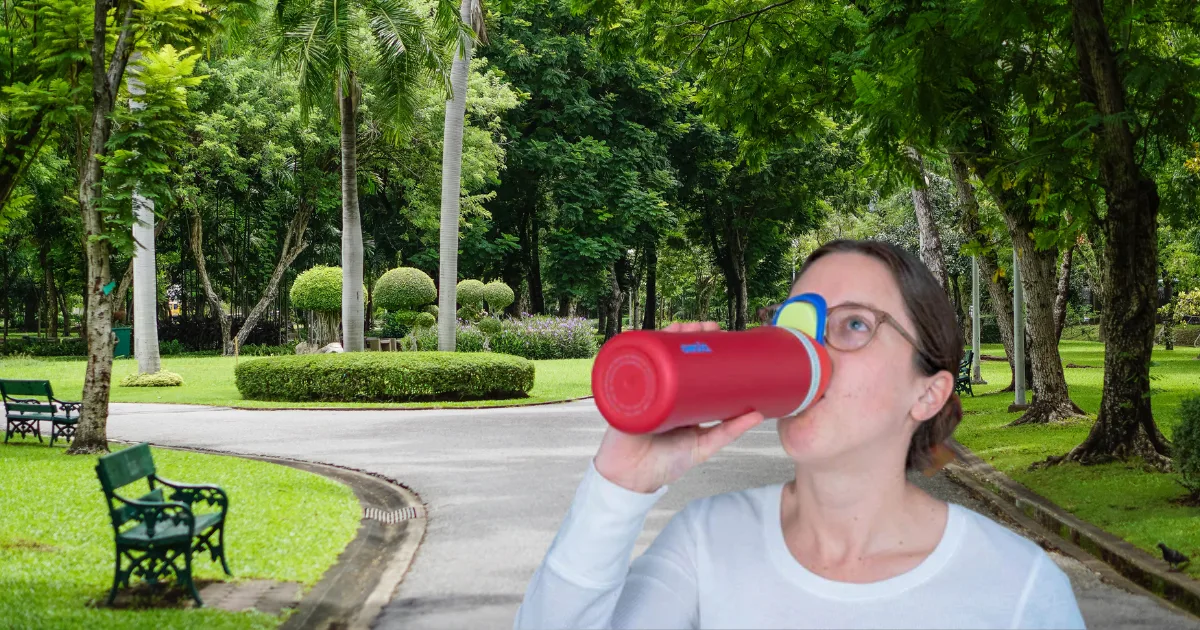 A person wearing glasses and a white shirt drinks from an Owala water bottle while standing on a path in a park. The park is lush with green trees, neatly trimmed bushes lining the walkway, and benches along the path—a perfect scene for embracing the ideal hydration solution.