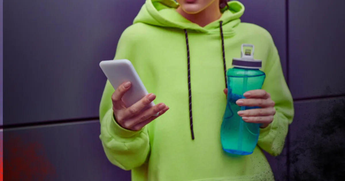 In a green hoodie, a woman holds a water bottle and a smartphone, showcasing a blend of fitness and technology.