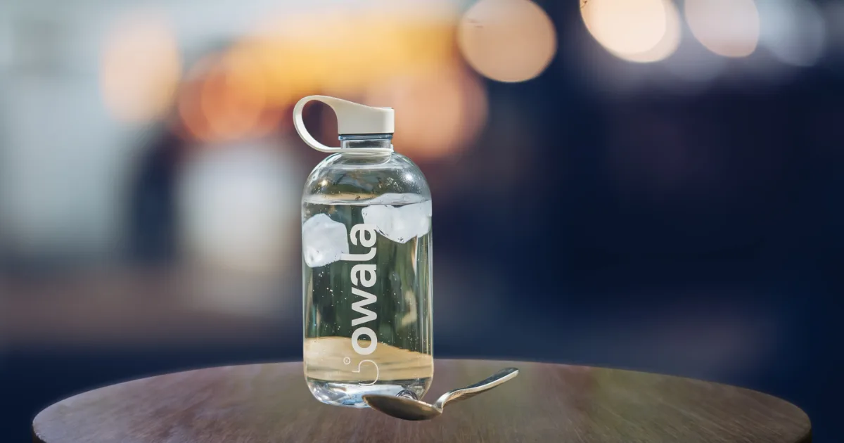 An Owala water bottle—a premium hydration solution—with ice cubes inside sits on a round wooden table. A metal spoon rests before it, while the softly blurred background with circular lights crafts a cozy atmosphere.
